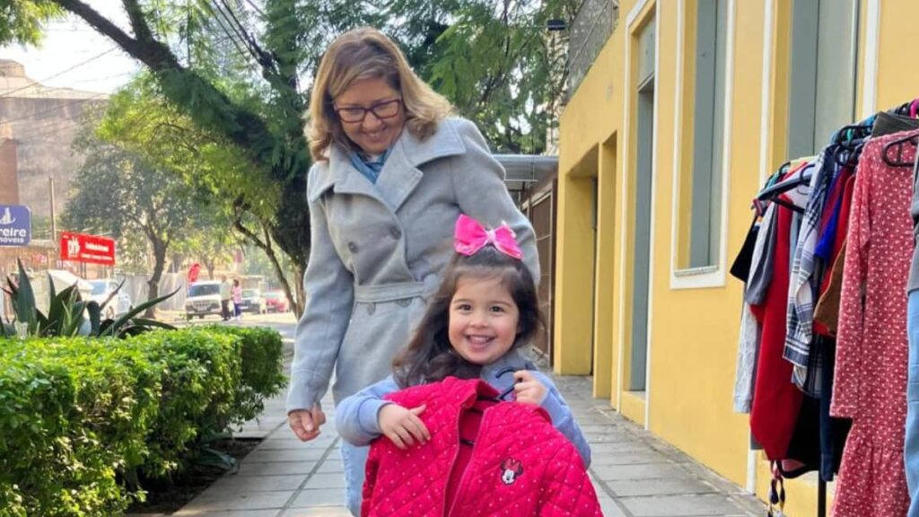 Criança sorridente ao lado de uma voluntária durante ação do brechó solidário do Instituto Alce, usando um casaco rosa da Minnie, em frente à antiga sede no bairro Menino Deus, em Porto Alegre.
