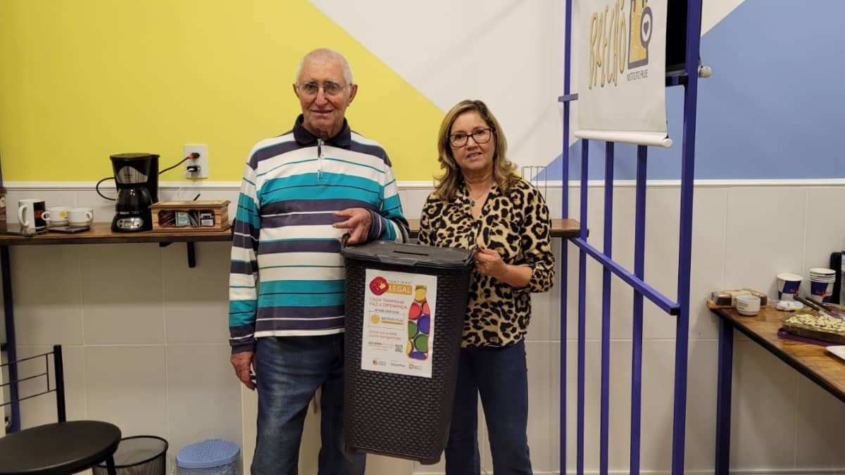 Homem e mulher em ambiente interno segurando coletor de tampinhas plásticas do programa Alce Tampinhas Solidárias.
