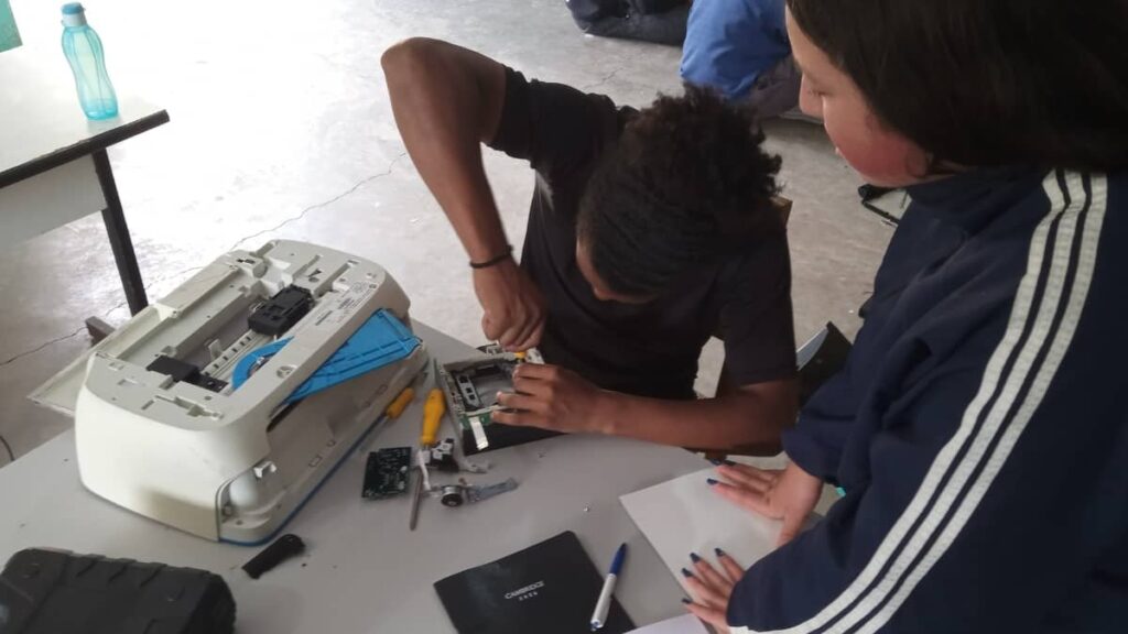 Dois participantes em oficina prática do Alce Tech trabalhando com equipamento eletrônico desmontado sobre uma mesa, com ferramentas e peças espalhadas ao redor.