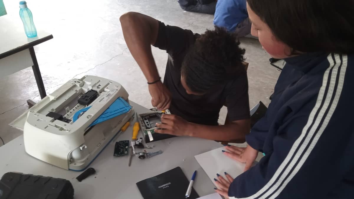 Dois participantes em oficina prática do Alce Tech trabalhando com equipamento eletrônico desmontado sobre uma mesa, com ferramentas e peças espalhadas ao redor.