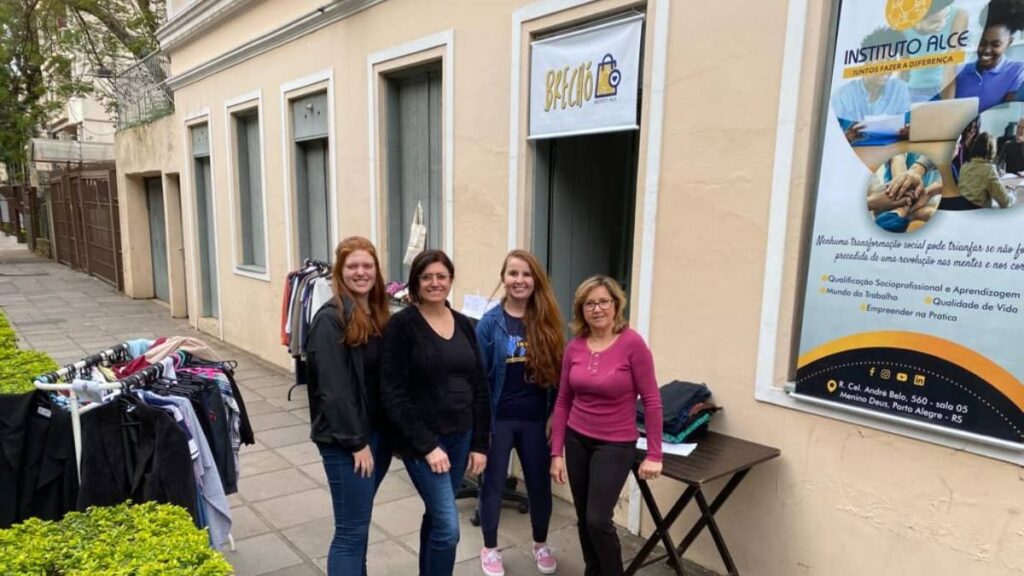 Quatro mulheres voluntárias em frente à antiga sede do Instituto Alce, no bairro Menino Deus, em Porto Alegre, durante uma das primeiras ações do brechó solidário, com araras de roupas ao lado.
