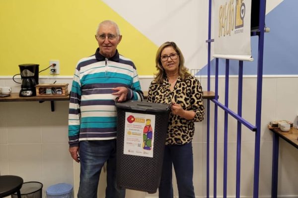 Homem e mulher em ambiente interno segurando coletor de tampinhas plásticas do programa Alce Tampinhas Solidárias.