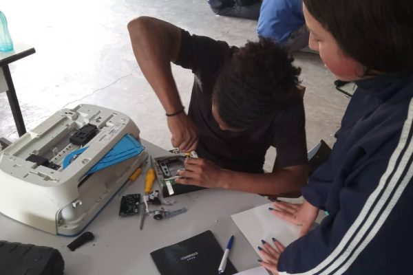 Dois participantes em oficina prática do Alce Tech trabalhando com equipamento eletrônico desmontado sobre uma mesa, com ferramentas e peças espalhadas ao redor.