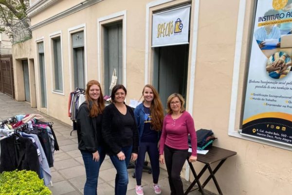 Quatro mulheres voluntárias em frente à antiga sede do Instituto Alce, no bairro Menino Deus, em Porto Alegre, durante uma das primeiras ações do brechó solidário, com araras de roupas ao lado.