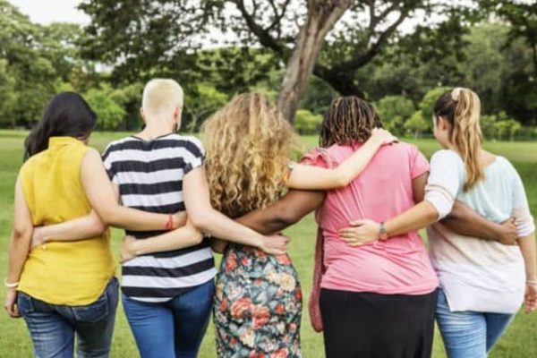 Quatro mulheres abraçadas de costas em parque, simbolizando união e empoderamento feminino no grupo de Terapia Comunitária.